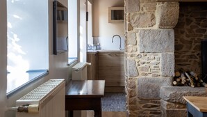 Un interior rústico con chimenea de piedra, un banco de madera, una zona de cocina con fregadero y un ventanal que proporciona luz natural.