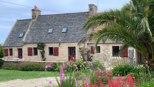 Una casa de piedra con techo inclinado, varias ventanas y un jardín con diversas flores.