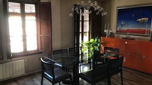 Un coin repas avec une table et des chaises en verre, un lustre, une fenêtre avec des stores et un tableau au mur.