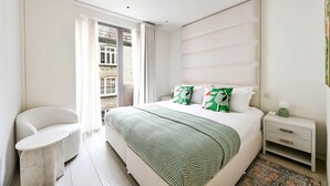 A modern bedroom with a large bed, a bedside table, a chair, and a view of the outside. - Luxe 3BR/4BA Home w/ Air Con Games 5min Selfridges (Greater London)