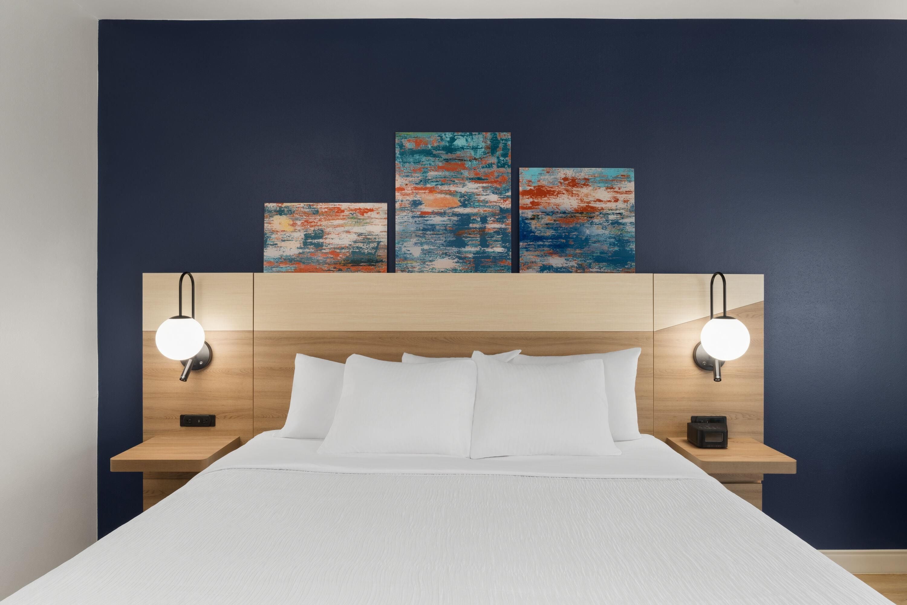 A hotel room with a bed, two bedside tables, a headboard with artwork, and wall-mounted reading lights.