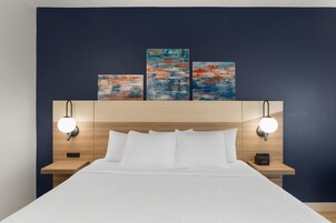 Una habitación de hotel con una cama, dos mesitas de noche, un cabecero decorado con obras de arte y luces de lectura fijadas a la pared.