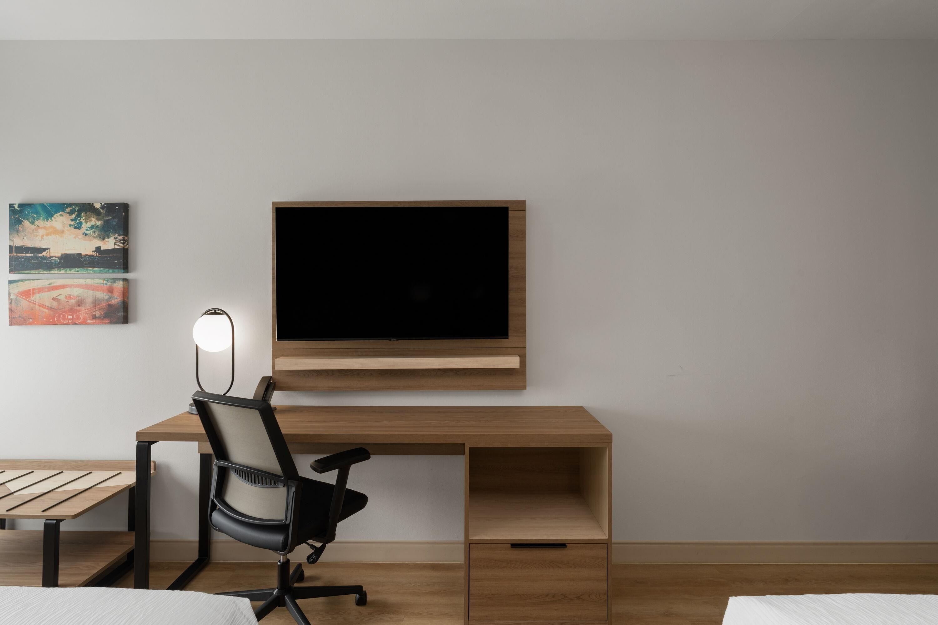 A modern room with a wooden desk, a black office chair, a flat-screen TV mounted on the wall, and a small table with a lamp.