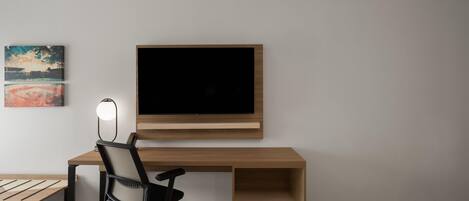 A modern room with a wooden desk, a black office chair, a flat-screen TV mounted on the wall, and a small table with a lamp.
