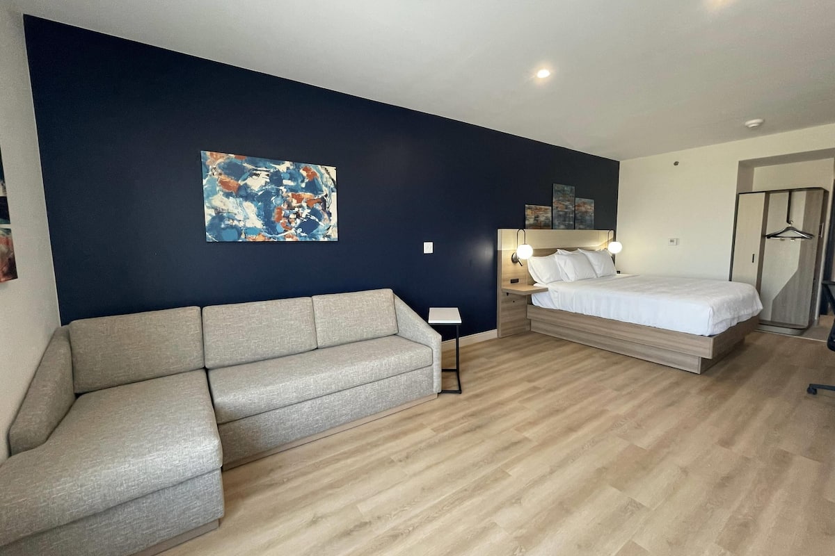 A modern hotel room with a grey sofa, a bed with white linens, a small bedside table, and a wall-mounted artwork.