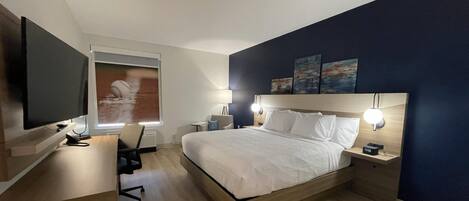 A modern hotel room with a bed, bedside tables, a desk, a chair, a TV, and artwork on the wall.