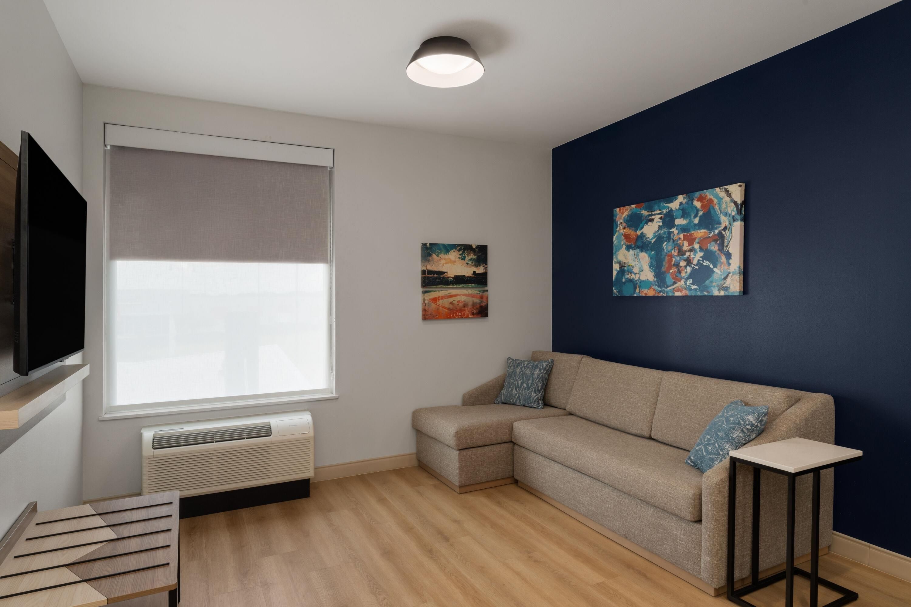 Una sala de estar moderna con un sofá color beige, una mesita, un televisor de pantalla plana, una ventana con persianas, un radiador y una pared azul decorada con un cuadro.