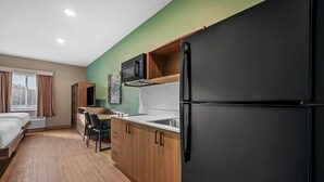 A compact hotel room with a kitchenette, a bed, a desk, and a window with curtains. - Echo Suites Extended Stay By Wyndham Naples (Naples)