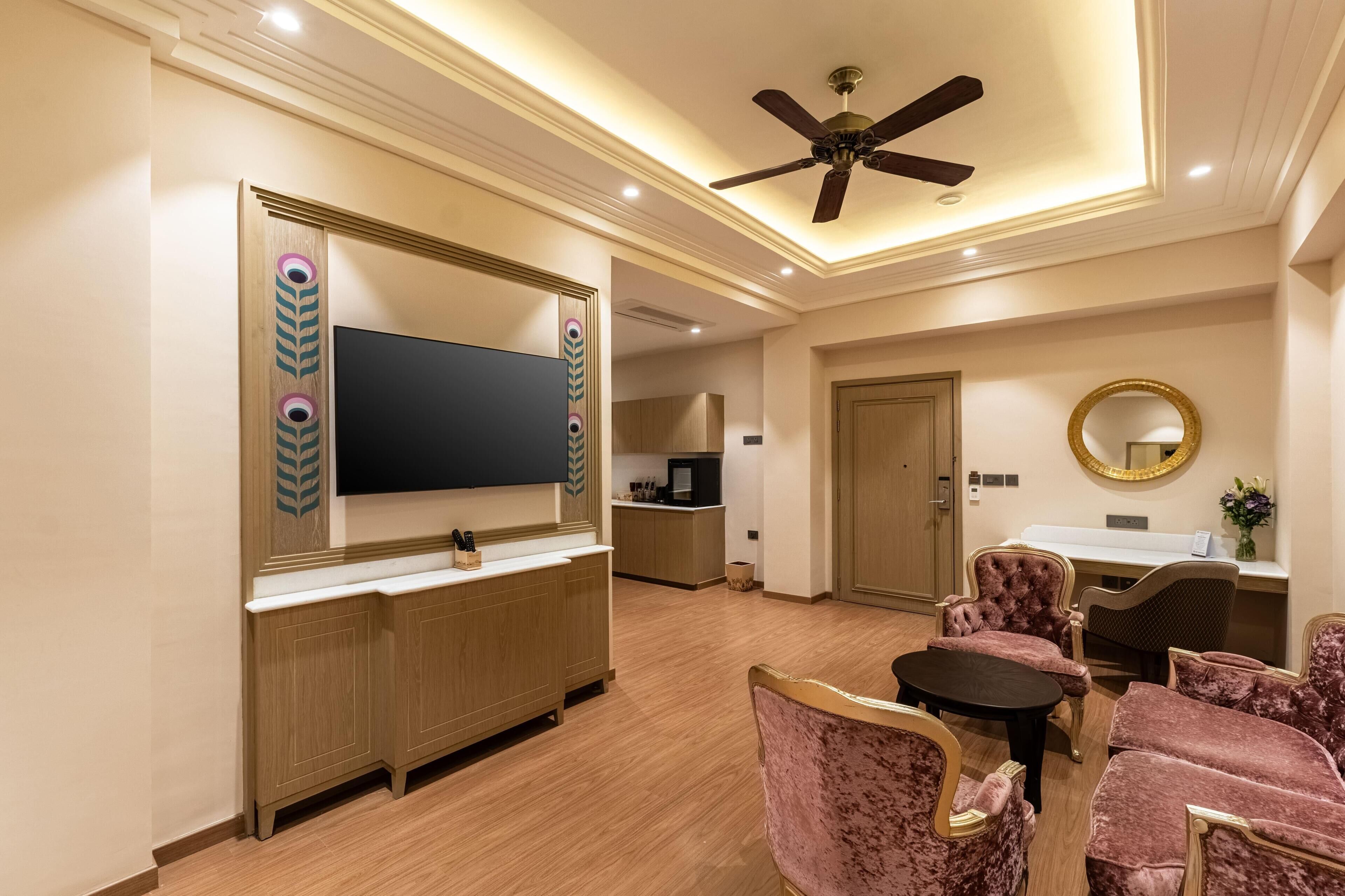A hotel room with a TV, a vanity with a mirror, a ceiling fan, and a dining area with chairs.