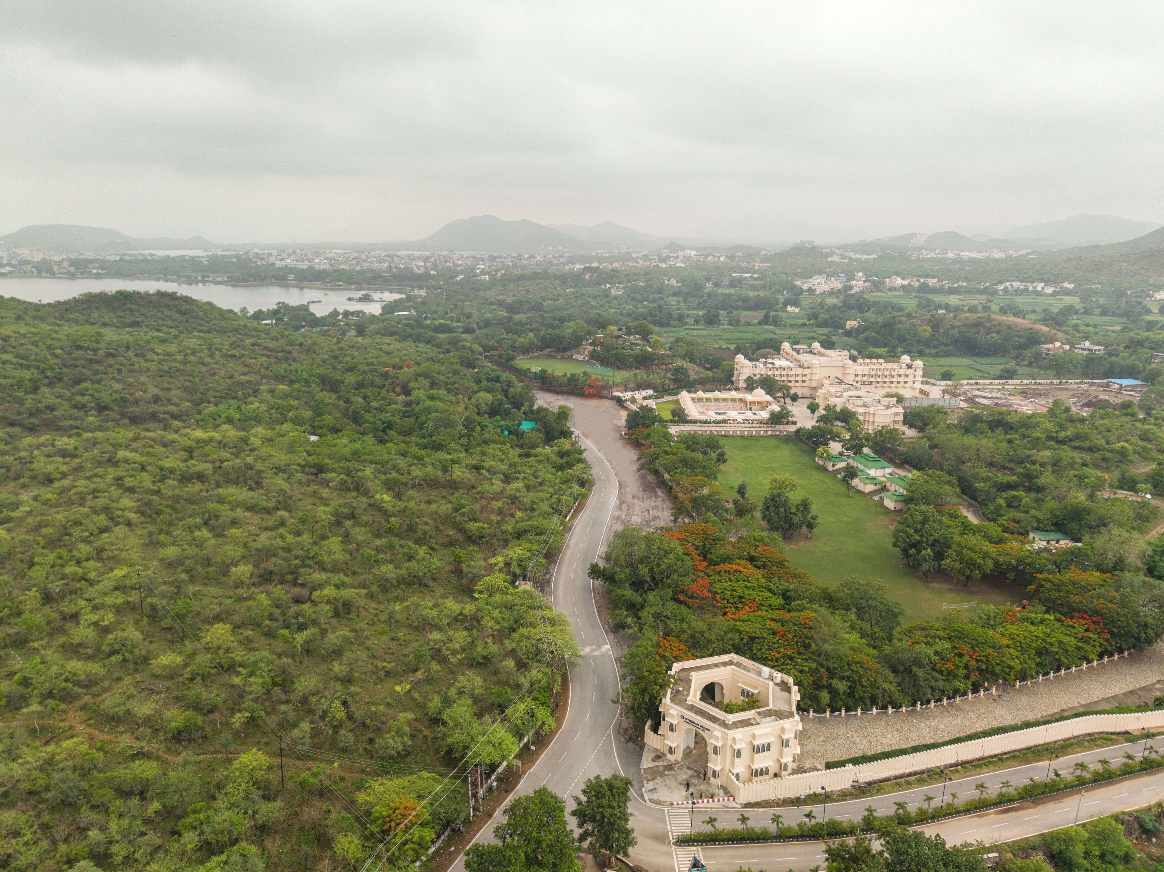 An aerial view of a large, historic building complex with multiple structures, surrounded by lush greenery and a winding road.