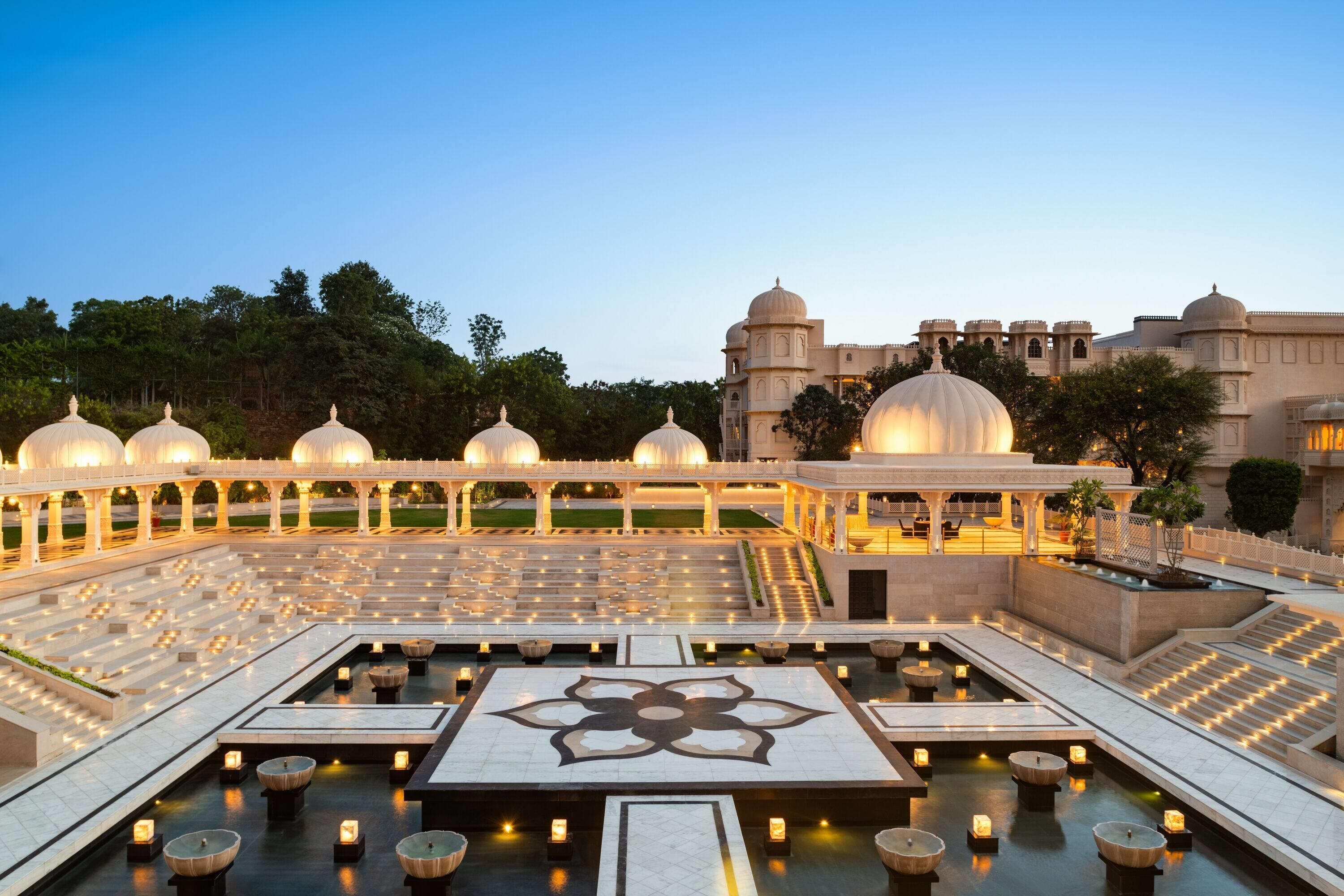 A courtyard with a central water feature, seating areas, and a large building with domed structures in the background.
