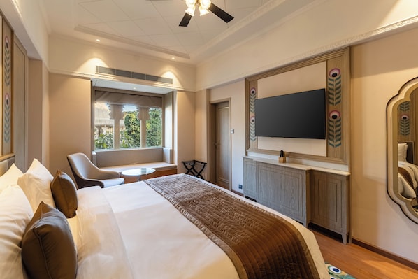 A hotel room with a bed, a TV, a desk, and a window with a view of greenery.