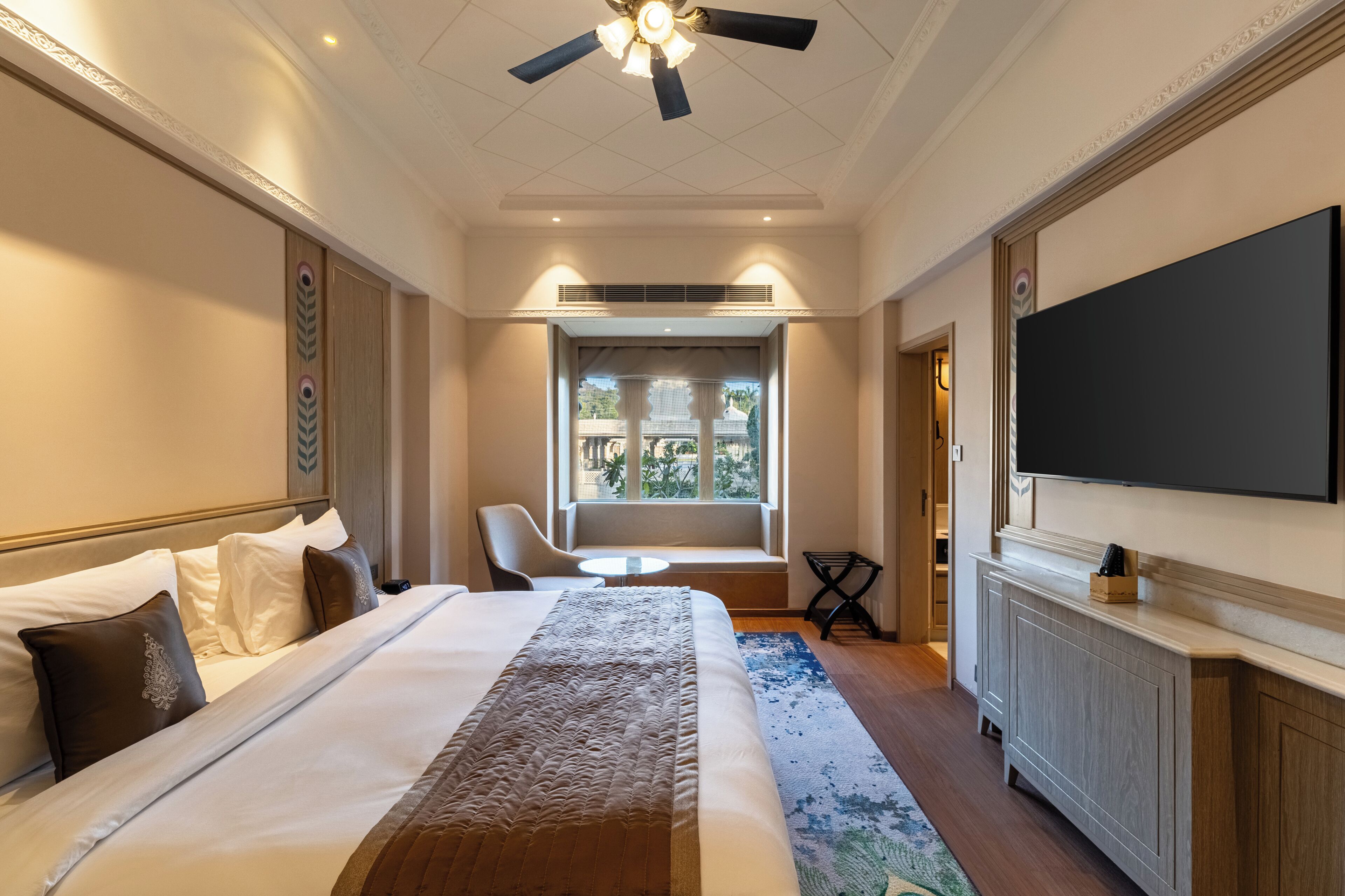A hotel room with a large bed, a TV, a ceiling fan, a window with a view, and a seating area by the window.