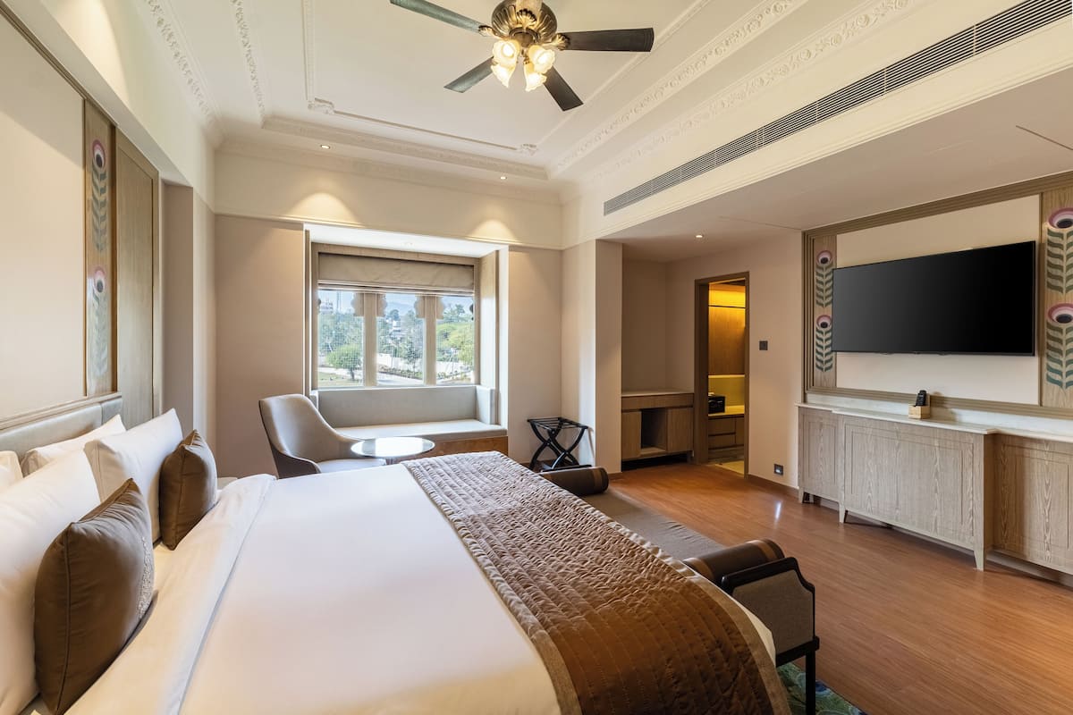 Ein Hotelzimmer mit einem großen Bett, einer Sitzecke inklusive Tisch, einem an der Wand montierten Fernseher und Blick ins Freie durch ein Fenster.