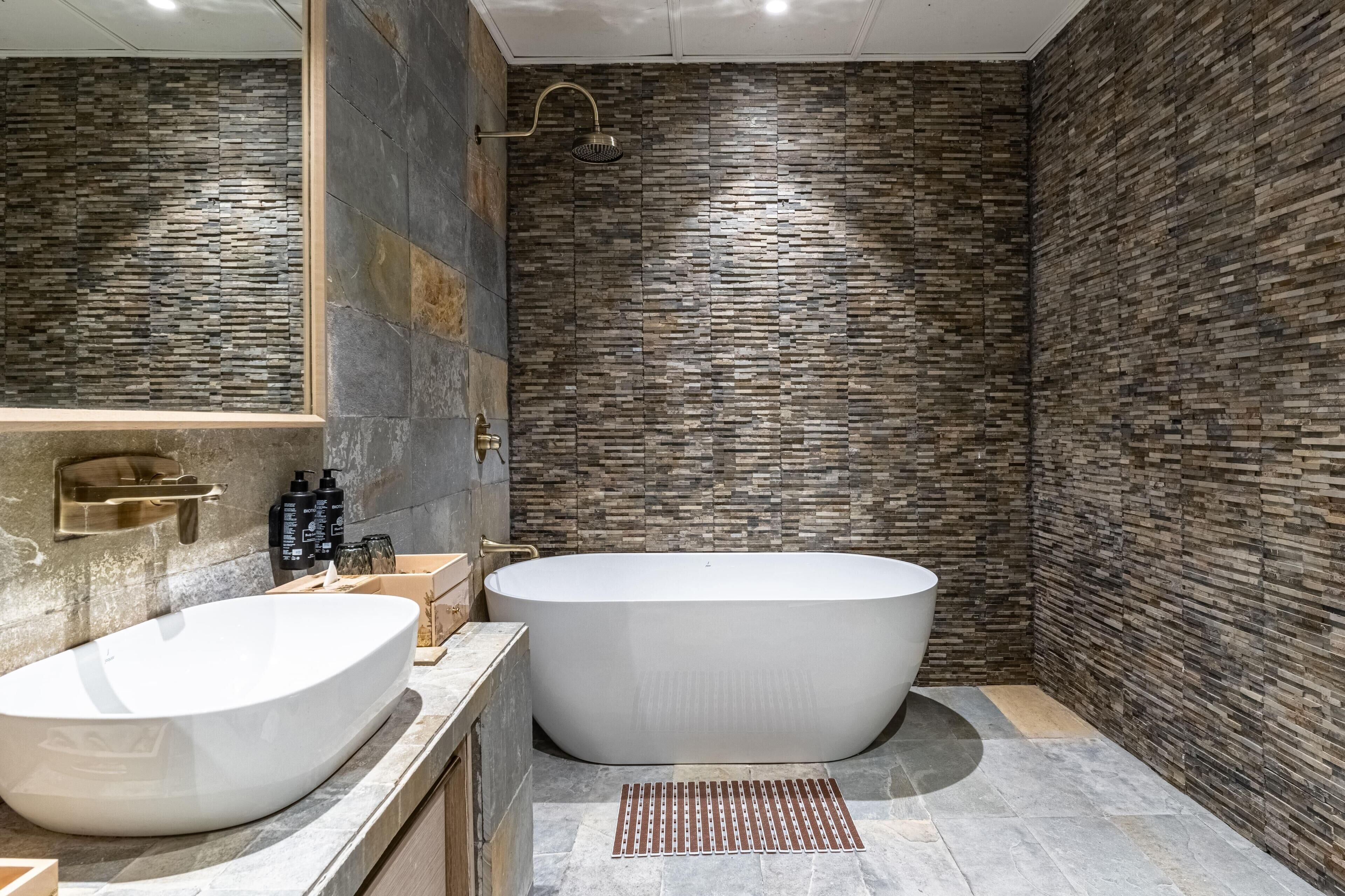A modern bathroom with a freestanding bathtub, a large mirror, and a stone-patterned wall.
