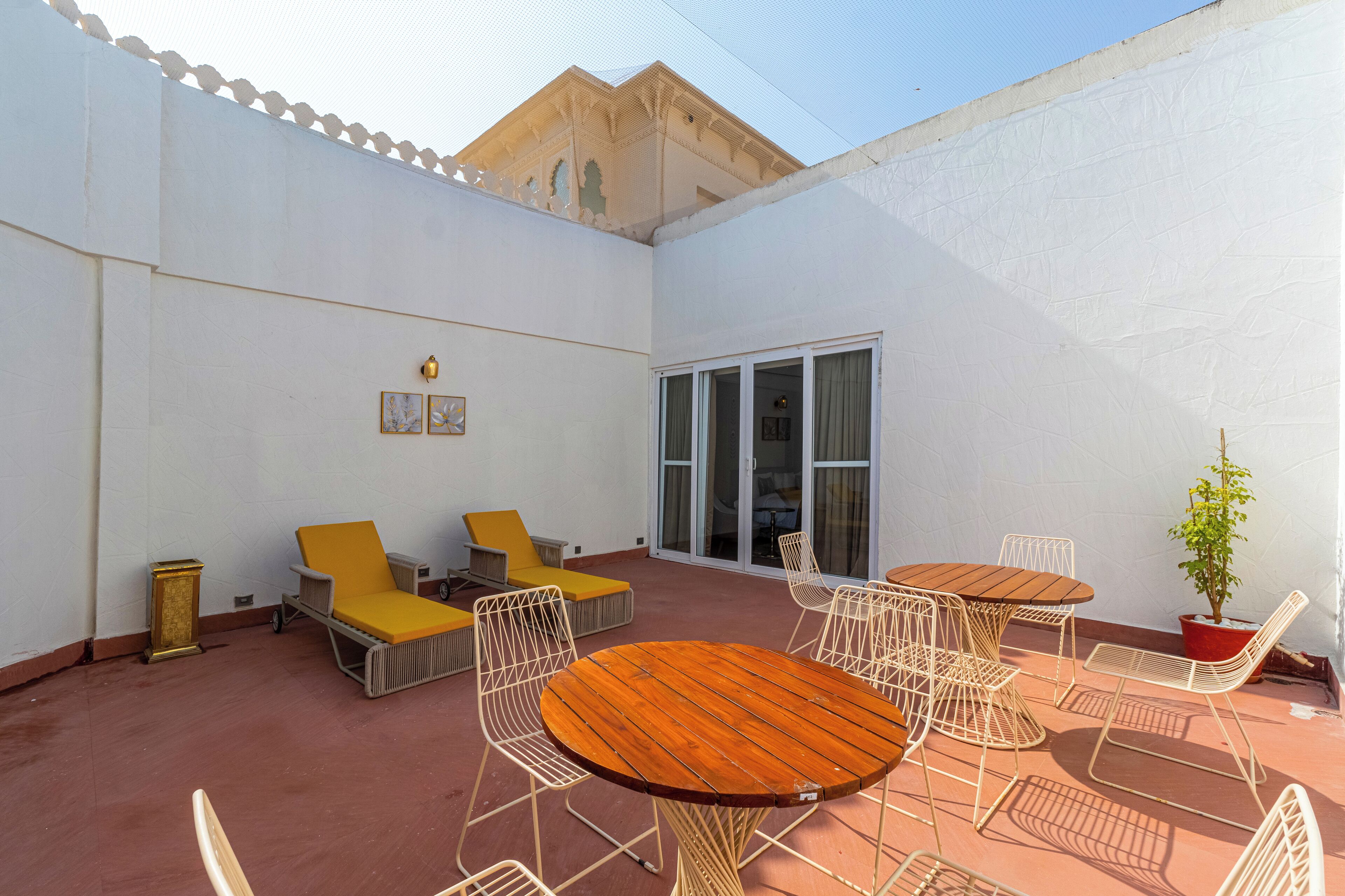 A sunlit patio with a round wooden table, white metal chairs, yellow lounge chairs, and a potted plant.