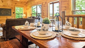 A wooden cabin interior with a dining table set for four, a sofa, and a large window with a view of trees. - Family Cabin, Private Indoor Pool, Hot Tub, Views, Arcades, EV - 5 Min to Pkwy (Pigeon Forge)