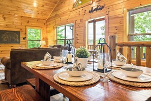 A wooden cabin interior with a dining table set for four, a sofa, and a large window with a view of trees. - Family Cabin w/ Indoor Pool, Hot Tub, Views & Arcades - 5 Min to Parkway (Pigeon Forge)