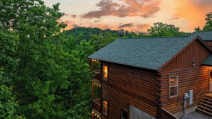 A wooden cabin with a balcony, surrounded by dense forest. - Family Cabin, Private Indoor Pool, Hot Tub, Views, Arcades, EV - 5 Min to Pkwy (Pigeon Forge)