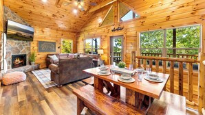 A wooden cabin interior with a fireplace, a dining table set for four, a sofa, and a large window overlooking greenery. - Family Cabin, Private Indoor Pool, Hot Tub, Views, Arcades, EV - 5 Min to Pkwy (Pigeon Forge)