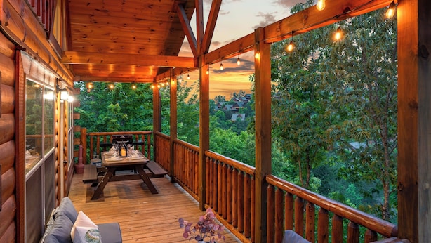 A wooden deck with a table set for dining, surrounded by wooden railings and a view of greenery. - Family Cabin, Private Indoor Pool, Hot Tub, Views, Arcades, EV - 5 Min to Pkwy (Pigeon Forge)