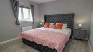 Une chambre à coucher avec une tête de lit capitonnée, un lit recouvert d’une couvre-lit à motifs, deux tables de chevet avec des lampes et une fenêtre avec des rideaux.