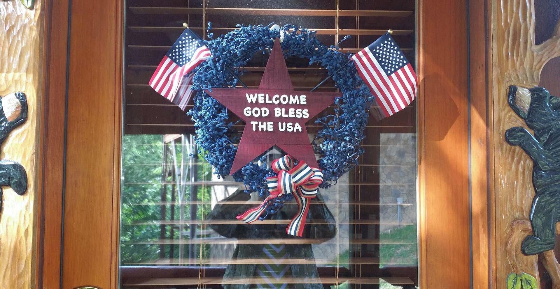 A wooden door with a star-shaped sign that reads 'WELCOME GOD BLESS THE USA' and two American flags.
