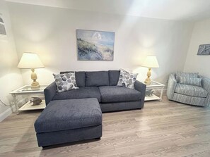 A living room with a grey sectional sofa, a matching ottoman, two side tables with lamps, a framed landscape painting, and a grey armchair.