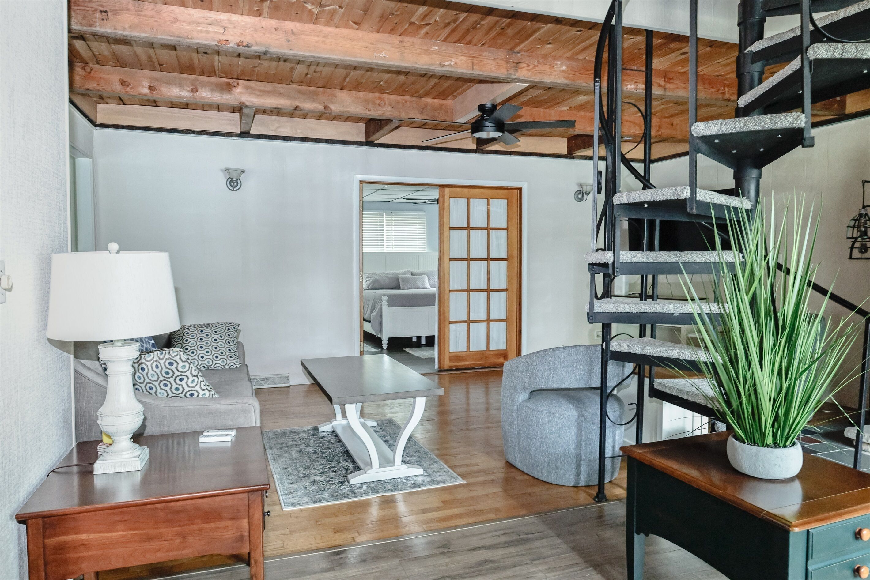 Een moderne woonkamer met een houten plafond, een wenteltrap, een grijze bank, een witte tafel en een potplant.