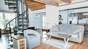 Un salon spacieux avec un escalier, un fauteuil gris, un canapé agrémenté de coussins à motifs et une table basse en bois.
