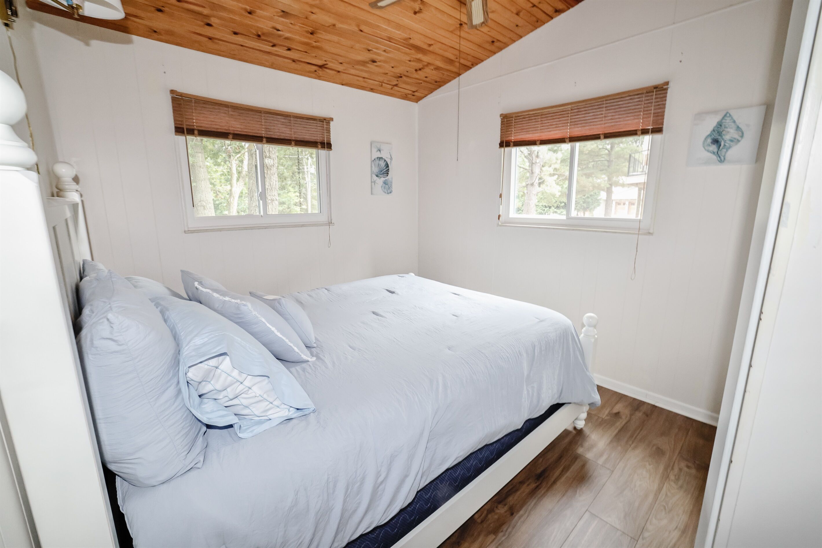 Ein Schlafzimmer mit Holzboden, Holzbalkendecke, zwei Fenstern mit Jalousien und einem Bett mit weißer Bettwäsche.