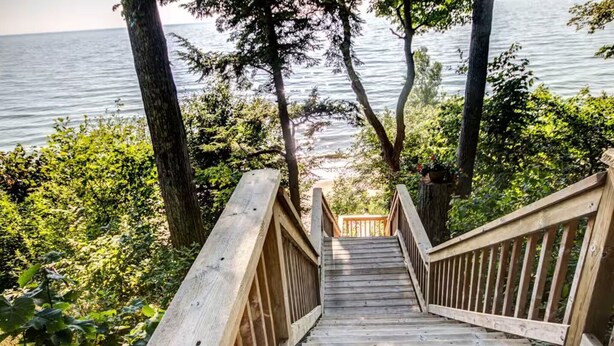 A wooden staircase leading to a dock by a lake, surrounded by trees and greenery. - Firepit, Deck & Views | Stairs to Lake Michigan (Montague)