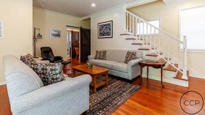 A living room with a sofa, armchair, and coffee table, a staircase with wooden railings, and a door leading to another room. - Firepit, Deck & Views | Stairs to Lake Michigan (Montague)