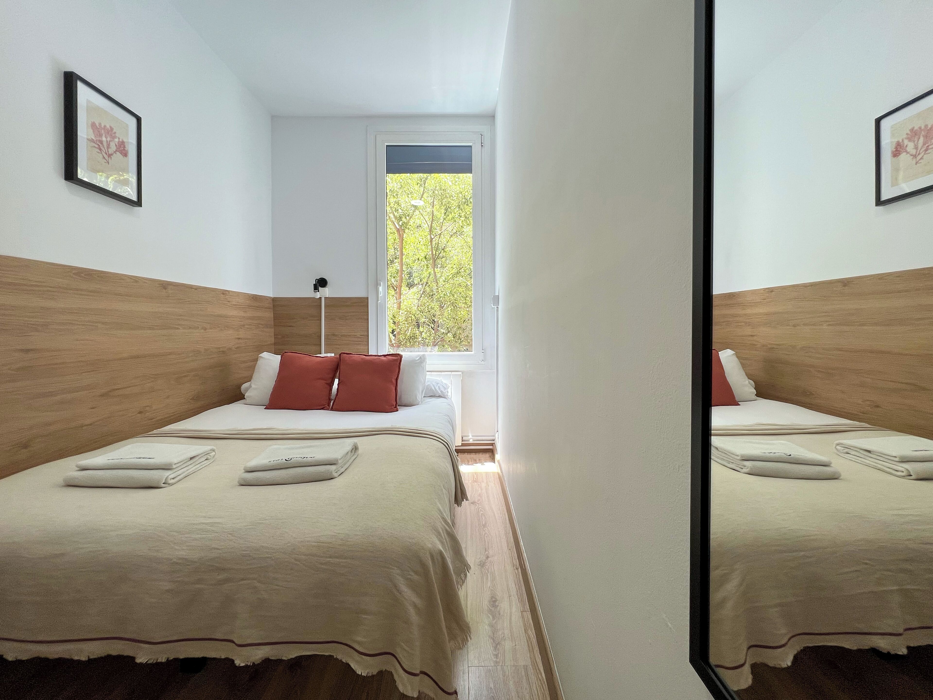 Ein Hotelzimmer mit zwei Betten, einem Fenster mit Blick auf Bäume und einem Spiegel, der den Raum reflektiert.