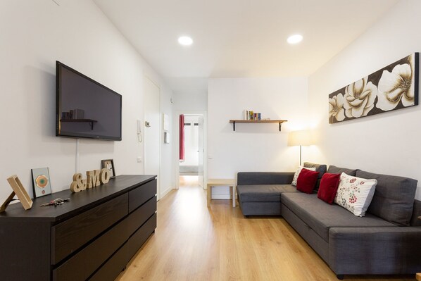 Un salon moderne avec un téléviseur à écran plat, une commode en bois foncé, un canapé gris avec des coussins rouges, une étagère en bois remplie de livres et un tableau floral accroché au mur.