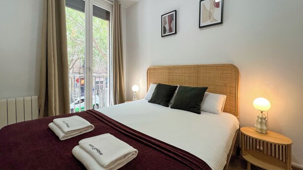 Een hotelkamer met een groot bed, twee bedlampen, een raam met gordijnen en twee ingelijste foto's aan de muur.