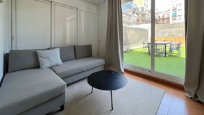 Een moderne woonkamer met een grijze bank, een klein zwart tafeltje en een grote glazen schuifdeur die uitkomt op een balkon met uitzicht op gebouwen en een hek.