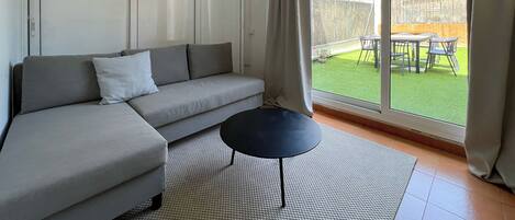A modern living room with a grey sofa, a small black table, and a large sliding glass door leading to a balcony with a view of buildings and a fence.