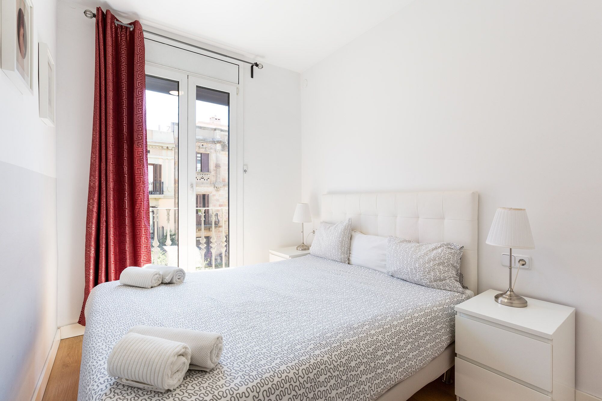 A bedroom with a bed, two bedside tables, a window with red curtains, and a view of buildings outside.