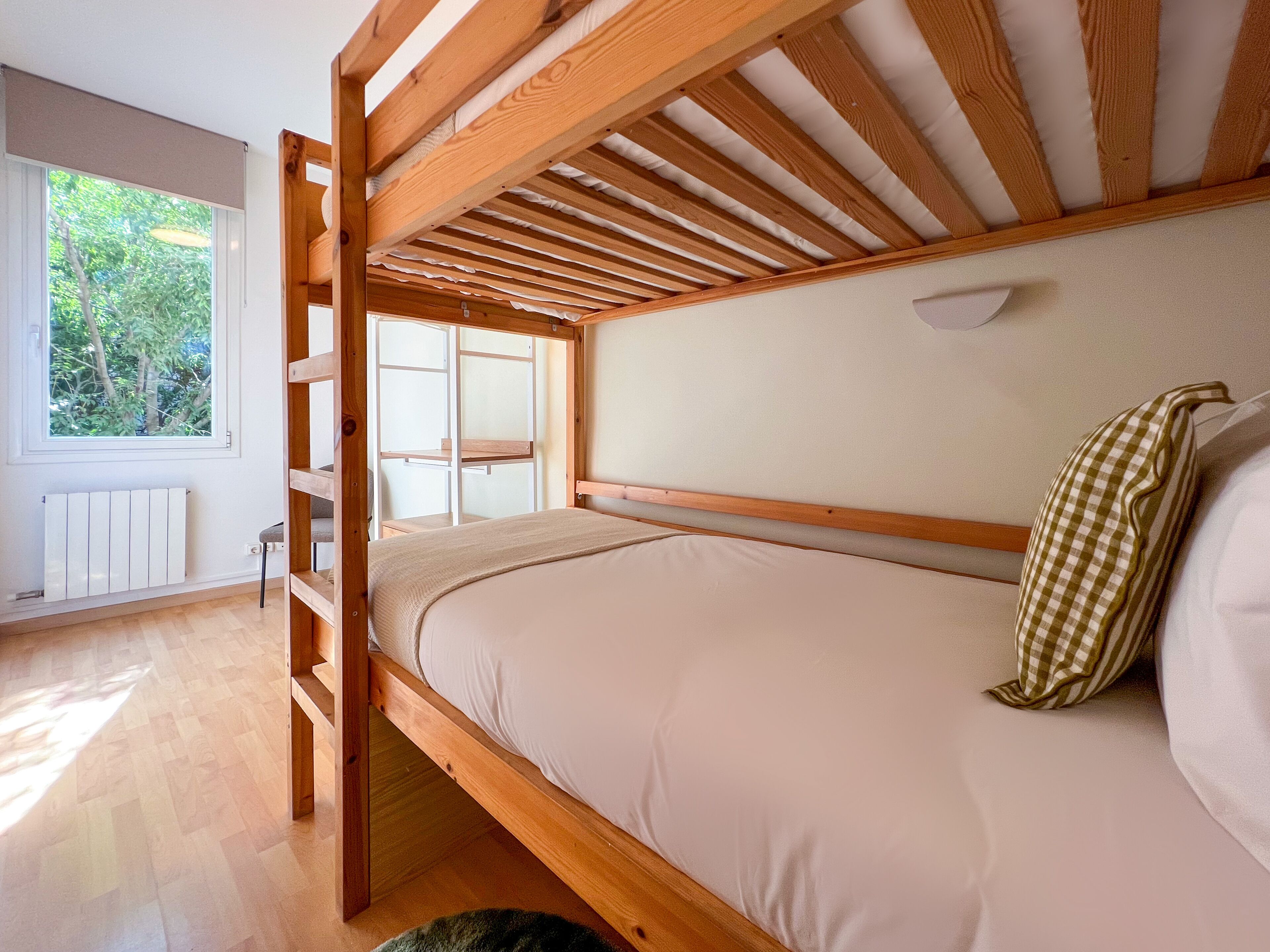 A bunk bed with a wooden frame and a white mattress. There is a window with a view of trees outside.