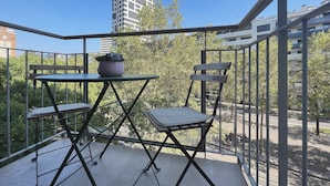 A balcony with a table and two chairs, a potted plant, and a view of a tall building and trees.