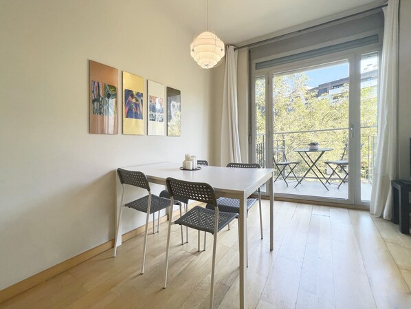 Une pièce moderne avec une table blanche et quatre chaises, un luminaire au plafond et un balcon aménagé avec du mobilier d’extérieur.