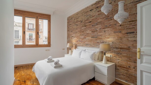 A bedroom with a brick wall, a large bed, two bedside tables with lamps, and a window with a view of buildings. - Stay U-nique Apartment Creu Coberta II (Barcelona)