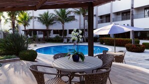 A poolside area with wicker chairs and a table, surrounded by palm trees and a building in the background. - HOTEL ORION (Itatiba)