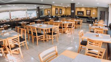 A spacious restaurant with multiple wooden tables and chairs, a counter with a beverage station, and framed artwork on the walls.