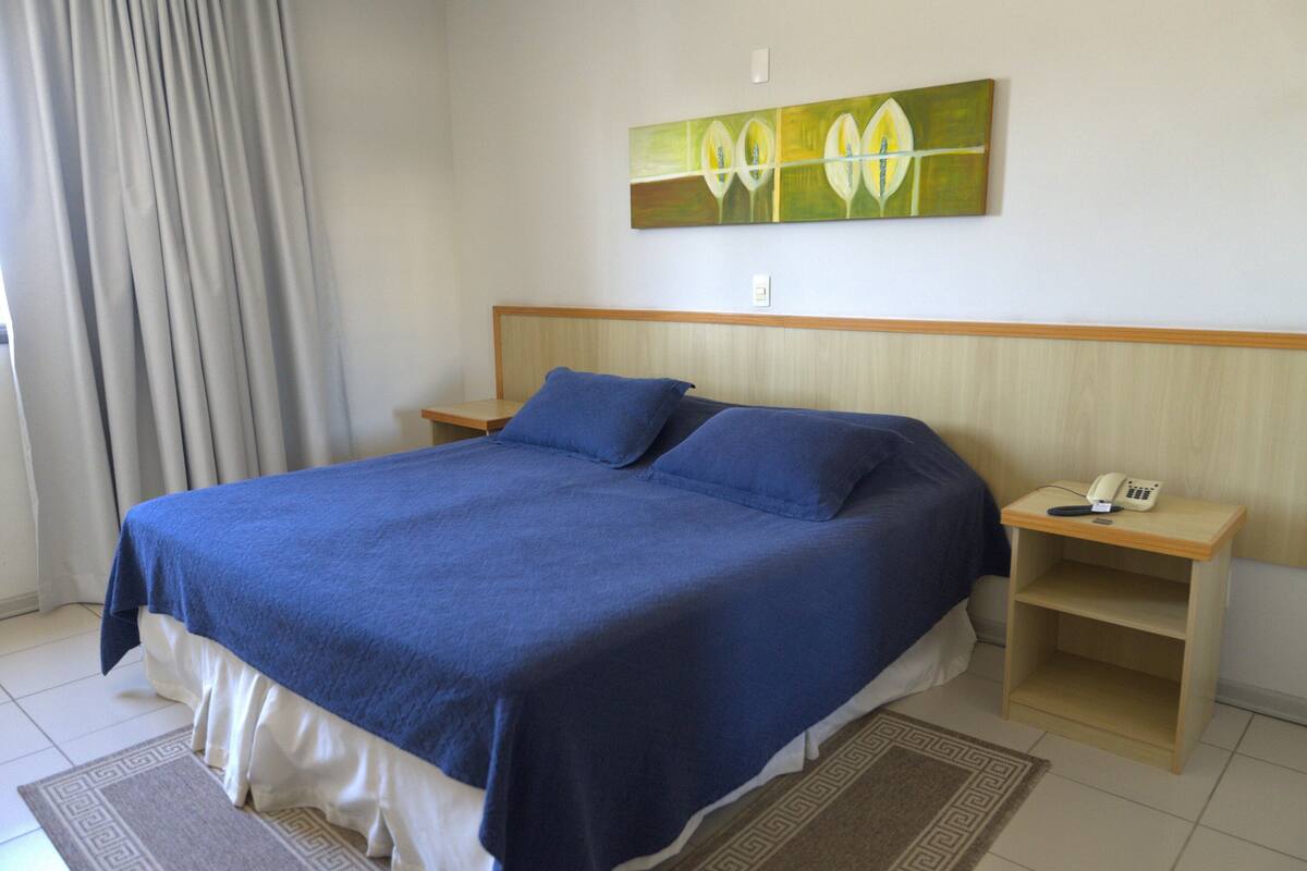 A neatly made bed with a blue bedspread and pillows, a bedside table with a telephone, and a painting on the wall.