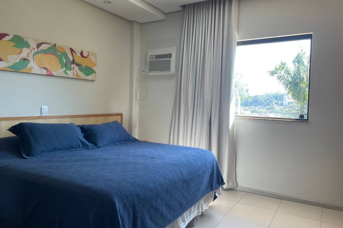 A bedroom with a bed, a window with curtains, a wall-mounted air conditioner, and a colorful abstract painting.