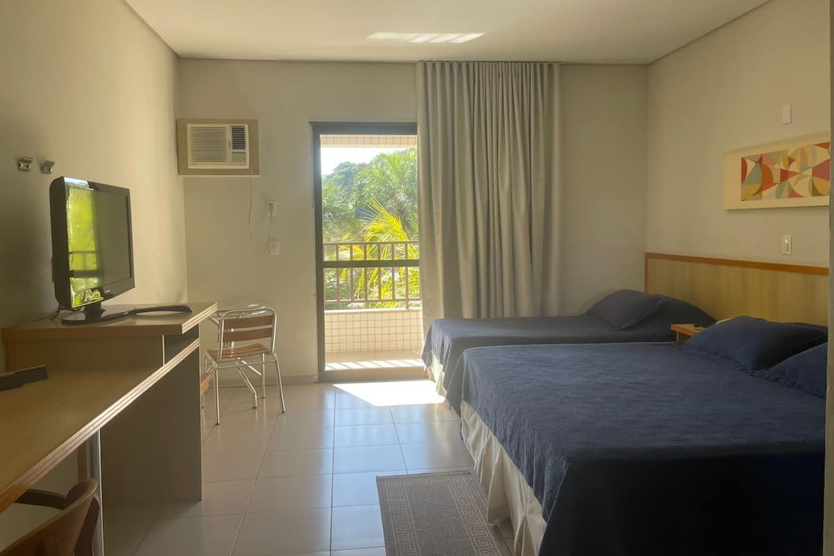 A hotel room with two beds, a desk with a computer, a chair, and a balcony with curtains.