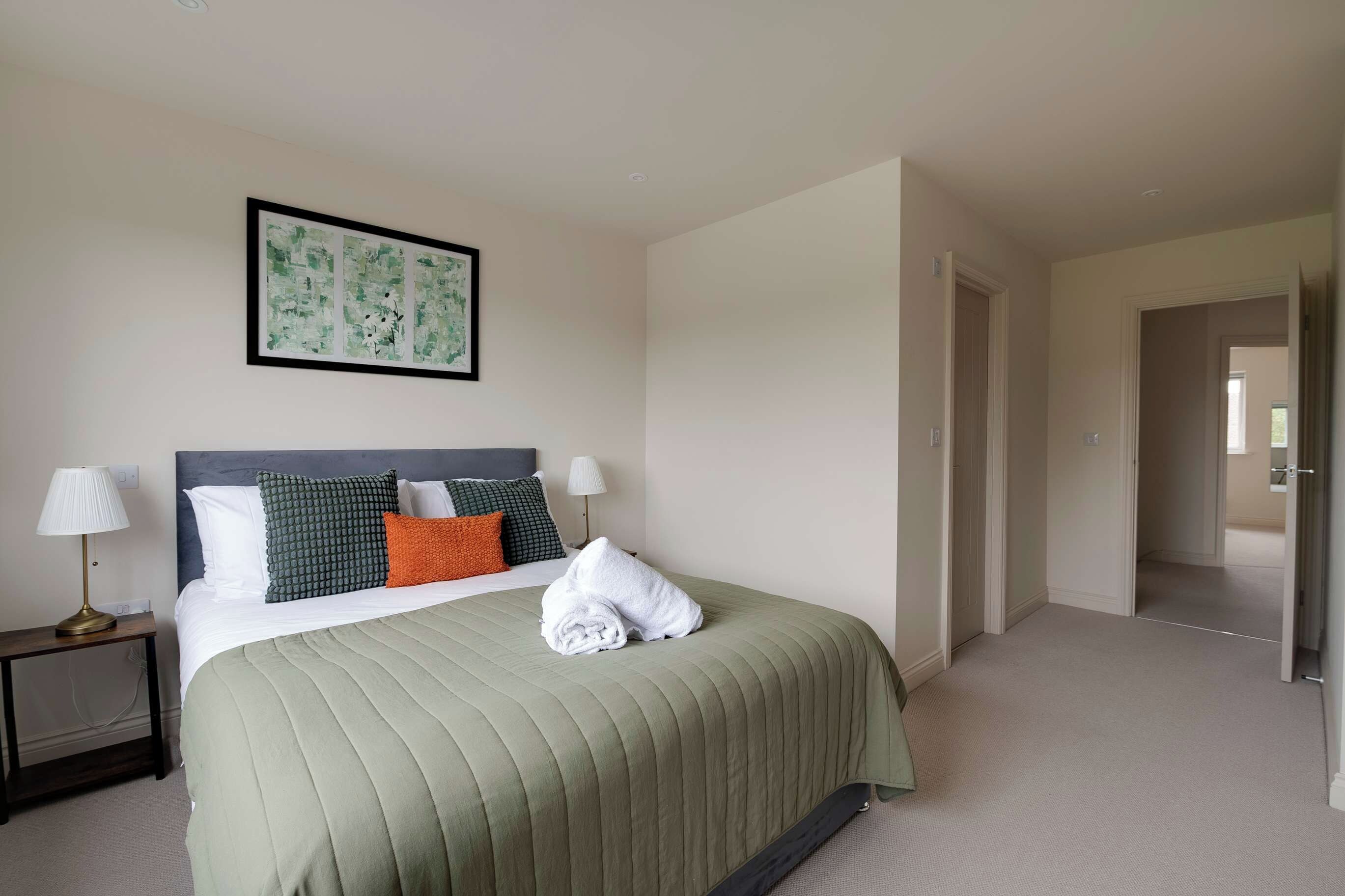 A hotel room with a bed, pillows, a framed artwork, and a nightstand with a lamp.