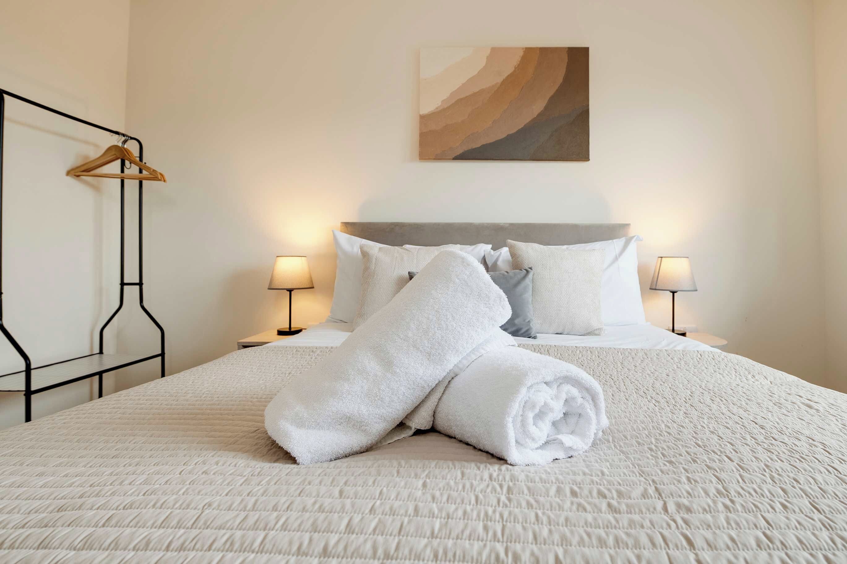 Un lit bien fait, avec du linge de lit blanc et des serviettes pliées, deux lampes de chevet, un cintre en bois et un tableau accroché au mur.
